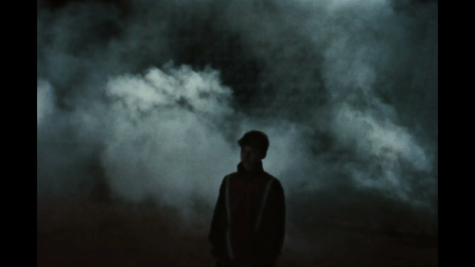 A young man in a dark jacket and cap stands alone in a smoky, dark environment, looking up.