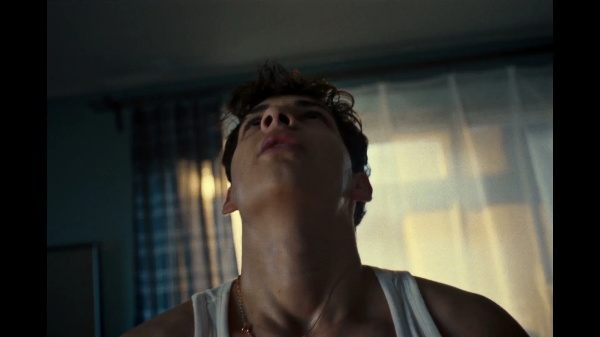 A young man in a white tank top looks up with a determined expression in a dimly lit room.