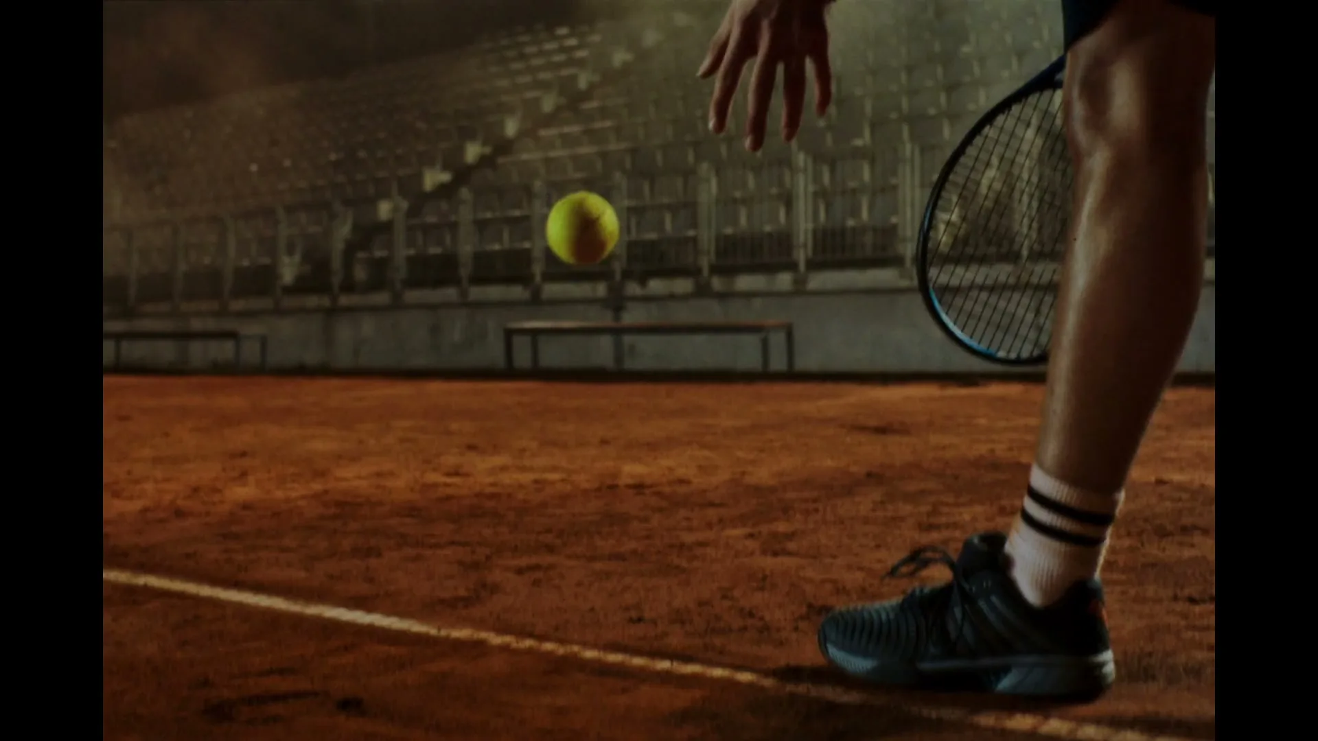 A tennis player in athletic wear bounces a tennis ball on a clay court in a stadium at night.