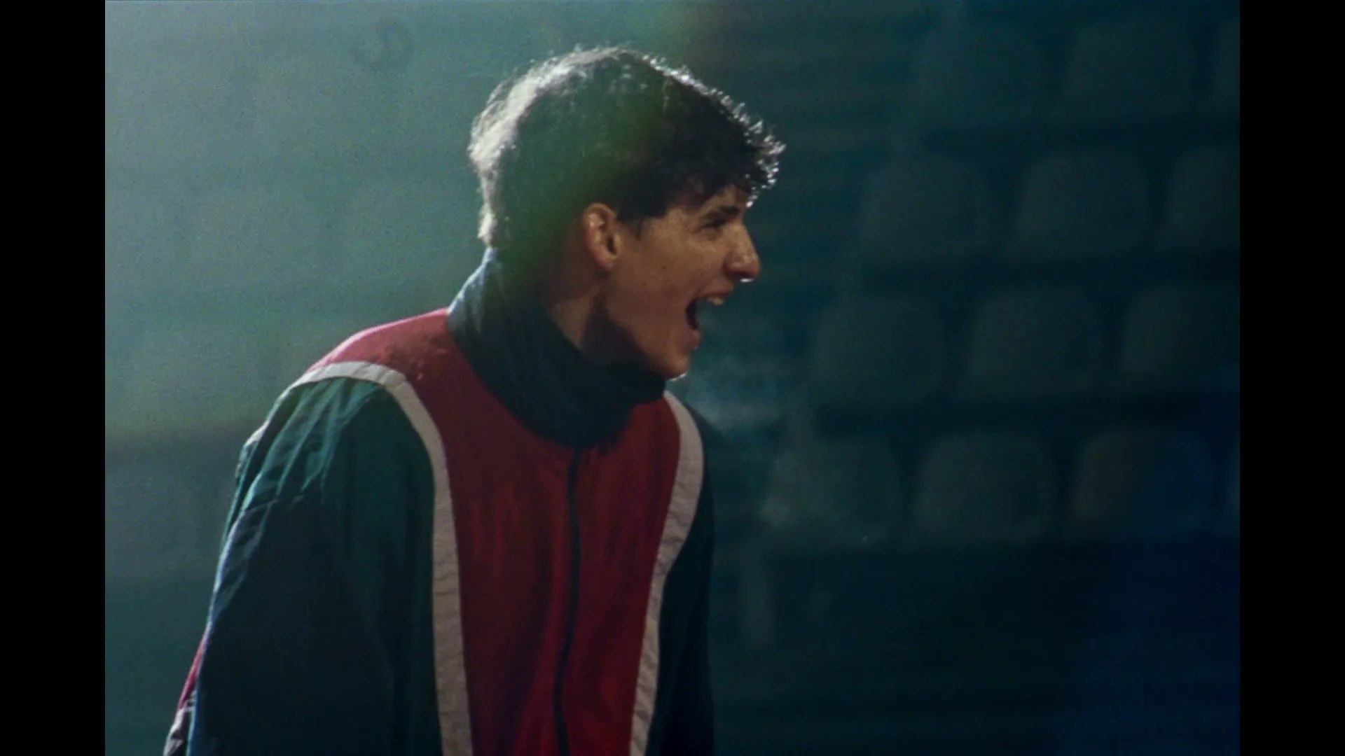 A young male athlete in a red and green tracksuit shouts with visible breath in a dark, empty stadium.
