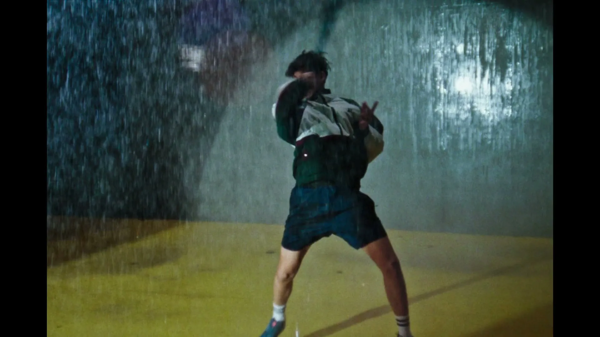A young male tennis player in a retro tracksuit hits a tennis ball on a wet, yellow court, with rain falling heavily around him.