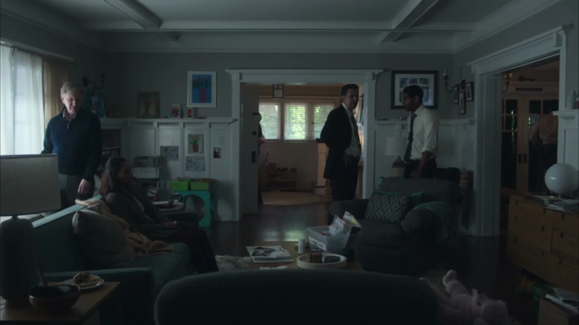A wide shot shows a family in their living room, with a man looking out the window, a woman sitting on a couch, and two men standing in the doorway.