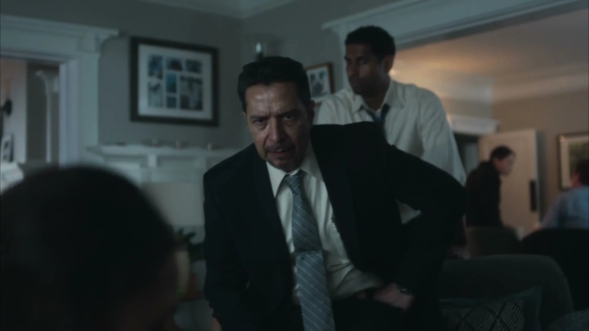 A male detective in a suit with a striped tie looks intently at someone off-screen, while other people are visible in the background of a living room.