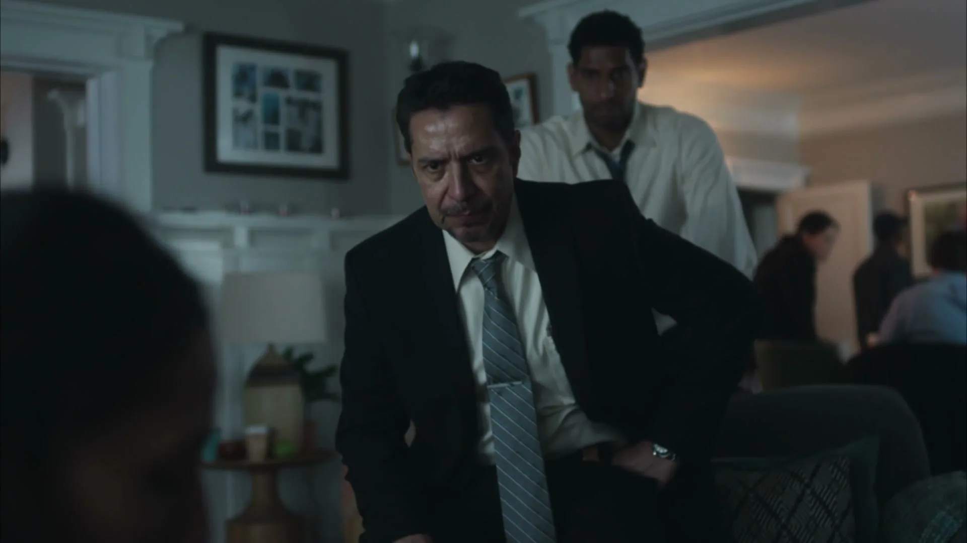 A man in a suit sits on a couch, looking concerned, while another man stands behind him in a dimly lit living room.
