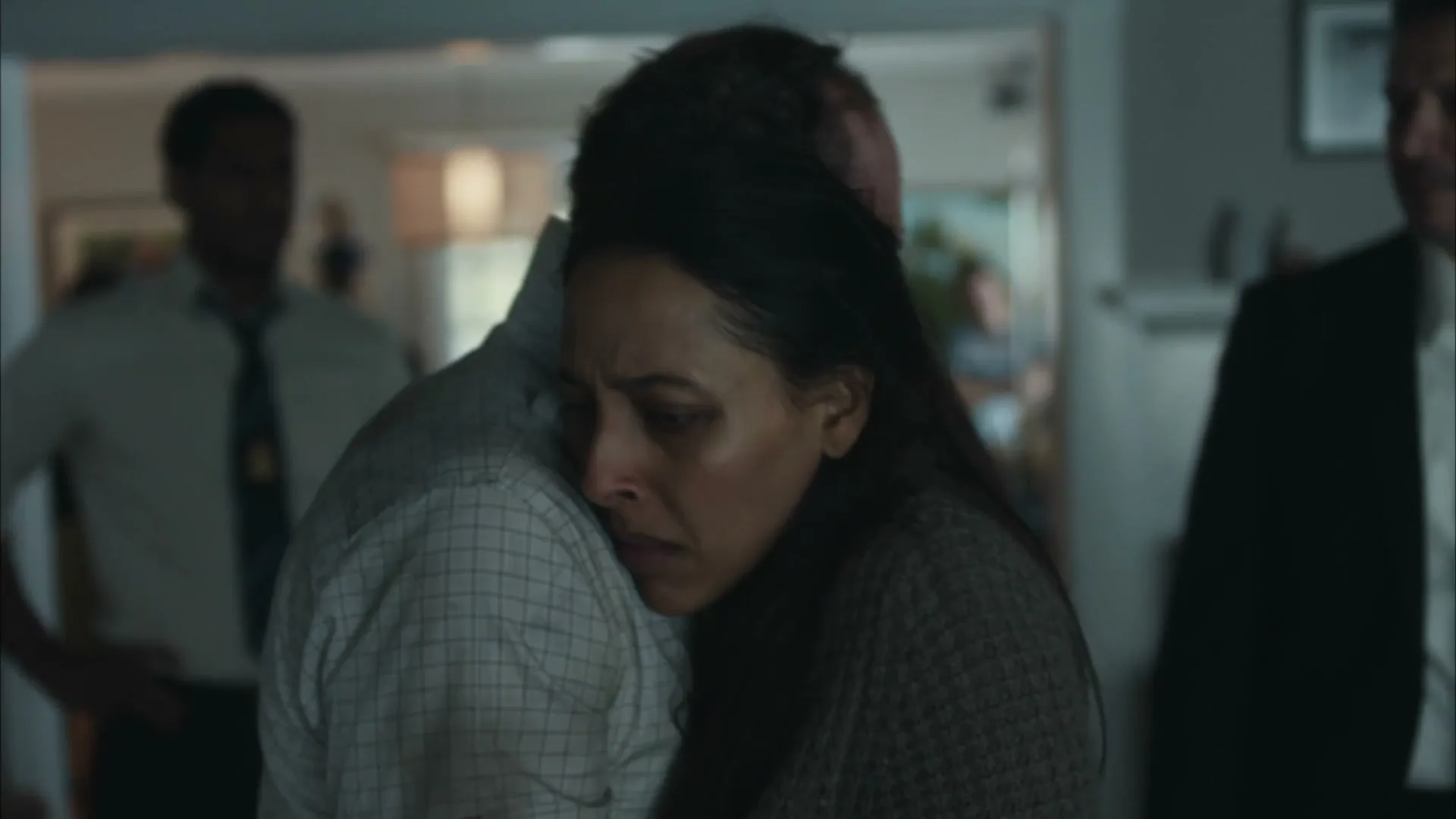 A woman with long dark hair, wearing a brown sweater, cries while hugging a man in a light-colored button-up shirt in a dimly lit house.
