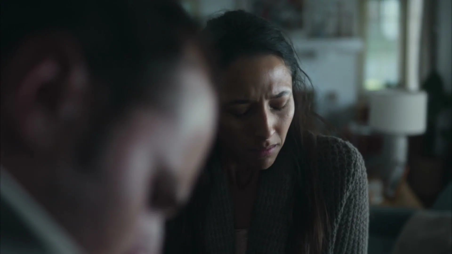 A woman with dark hair and a brown cardigan looks distressed and speaks to an unseen person in a dimly lit living room.
