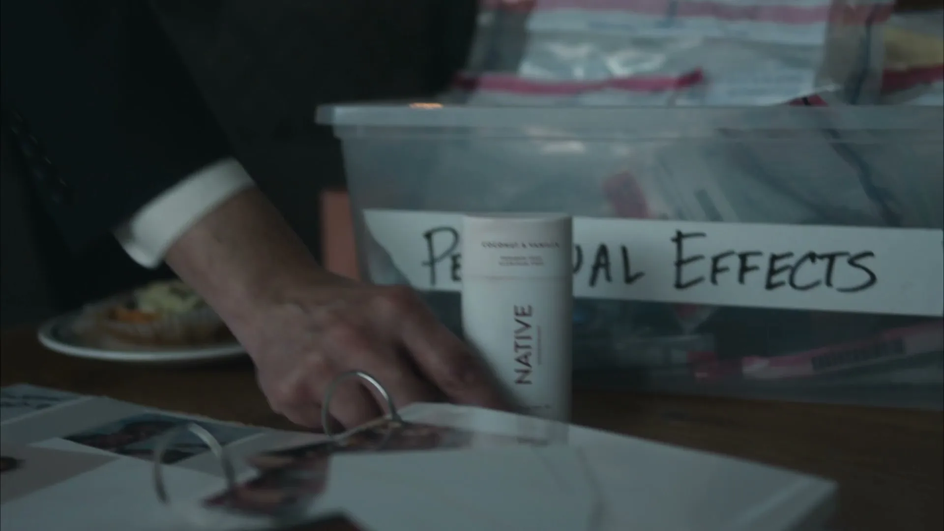 A man's hand in a suit reaches for a Native deodorant stick on a table next to a box labeled "Personal Effects" and a photo album.