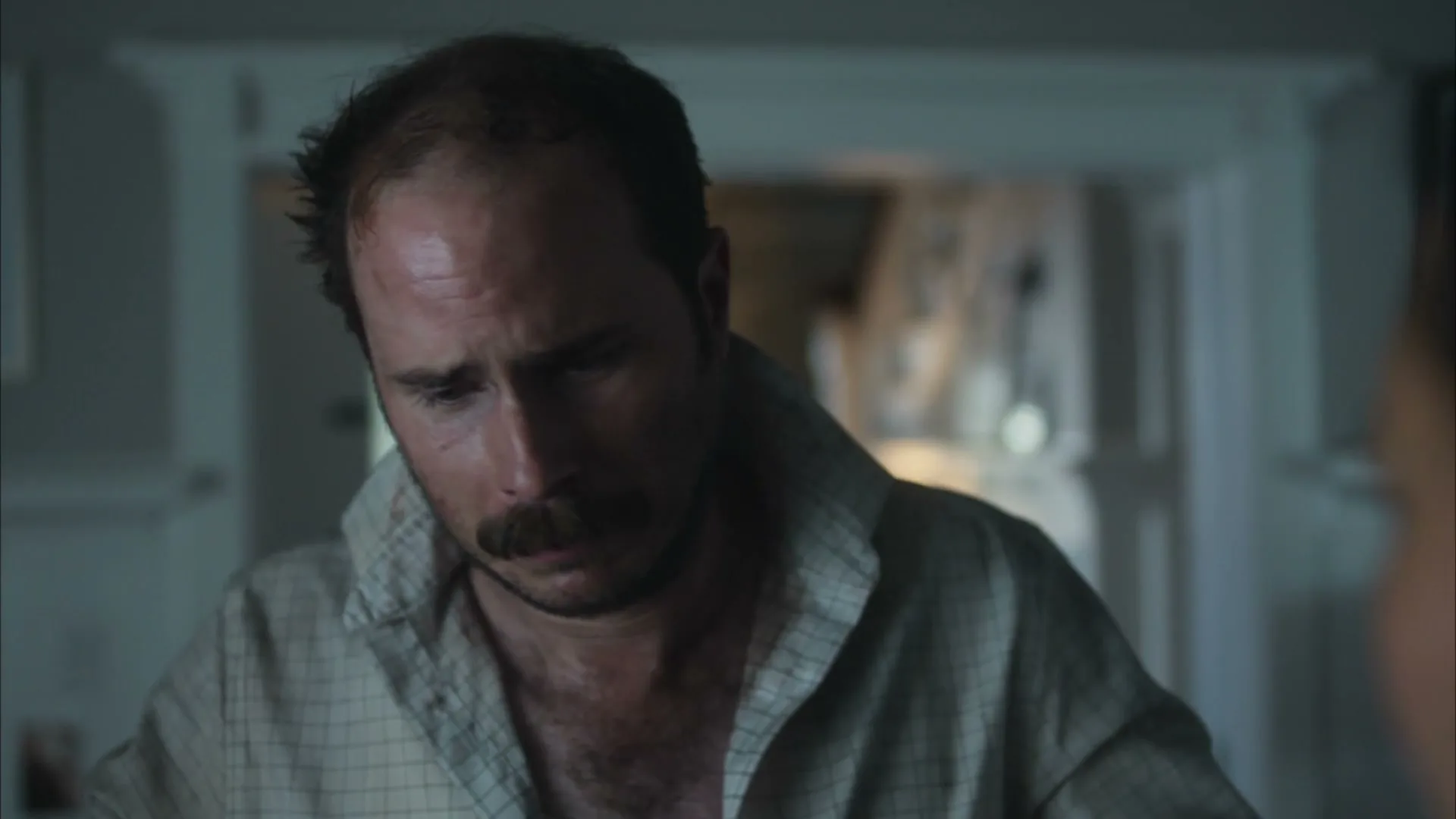A man with a mustache and receding hairline looks down, wearing an unbuttoned plaid shirt, in a dimly lit interior.