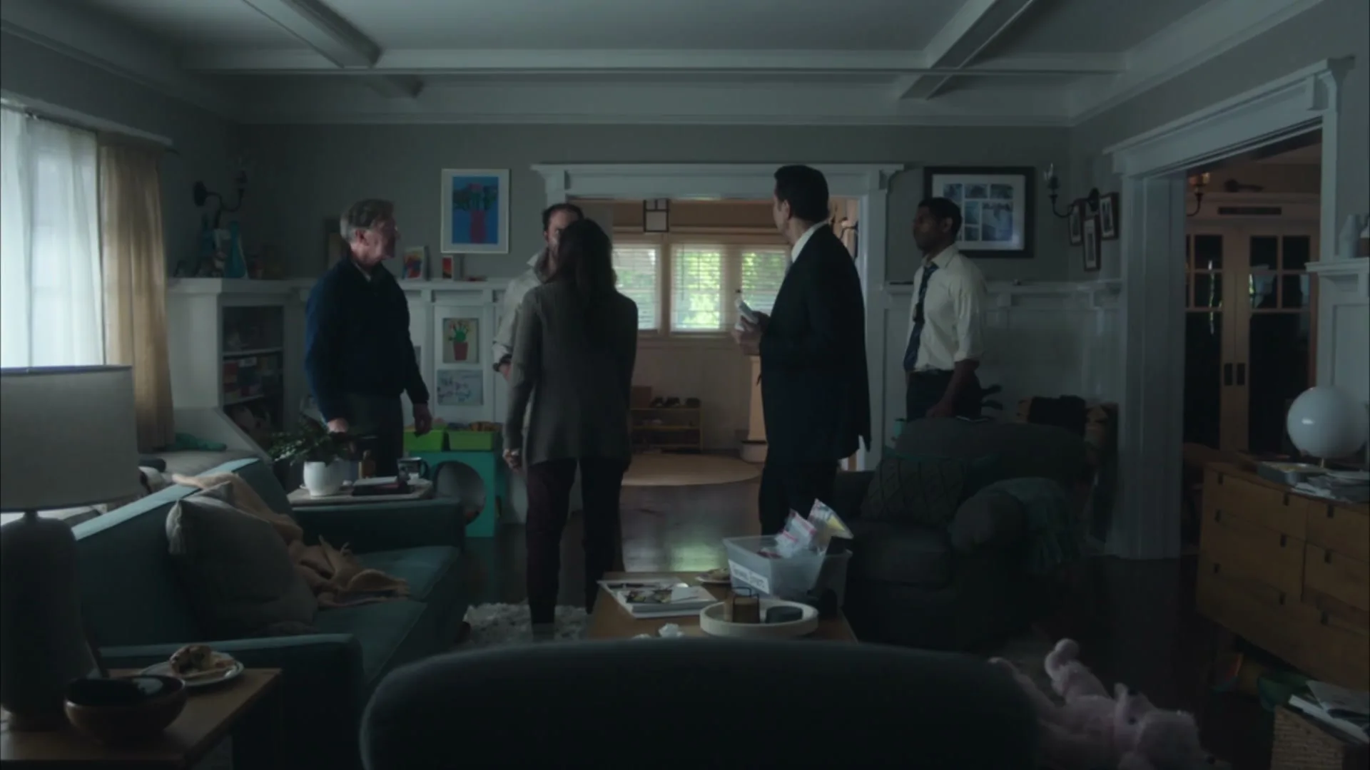 Five adults, three men and two women, stand in a dimly lit living room, talking amongst themselves.