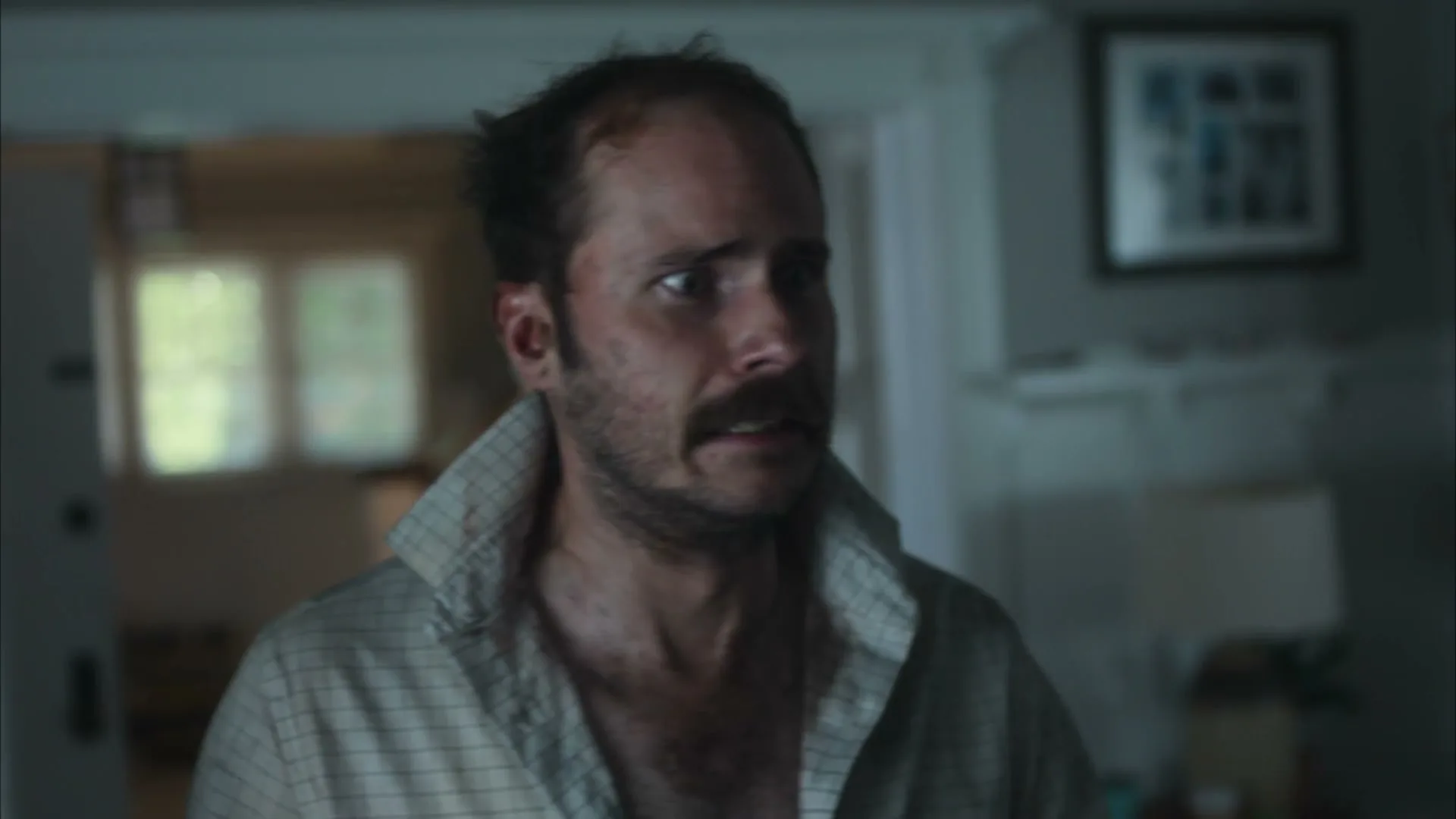A disheveled man with a mustache and an open shirt looks around nervously in a dimly lit house interior.