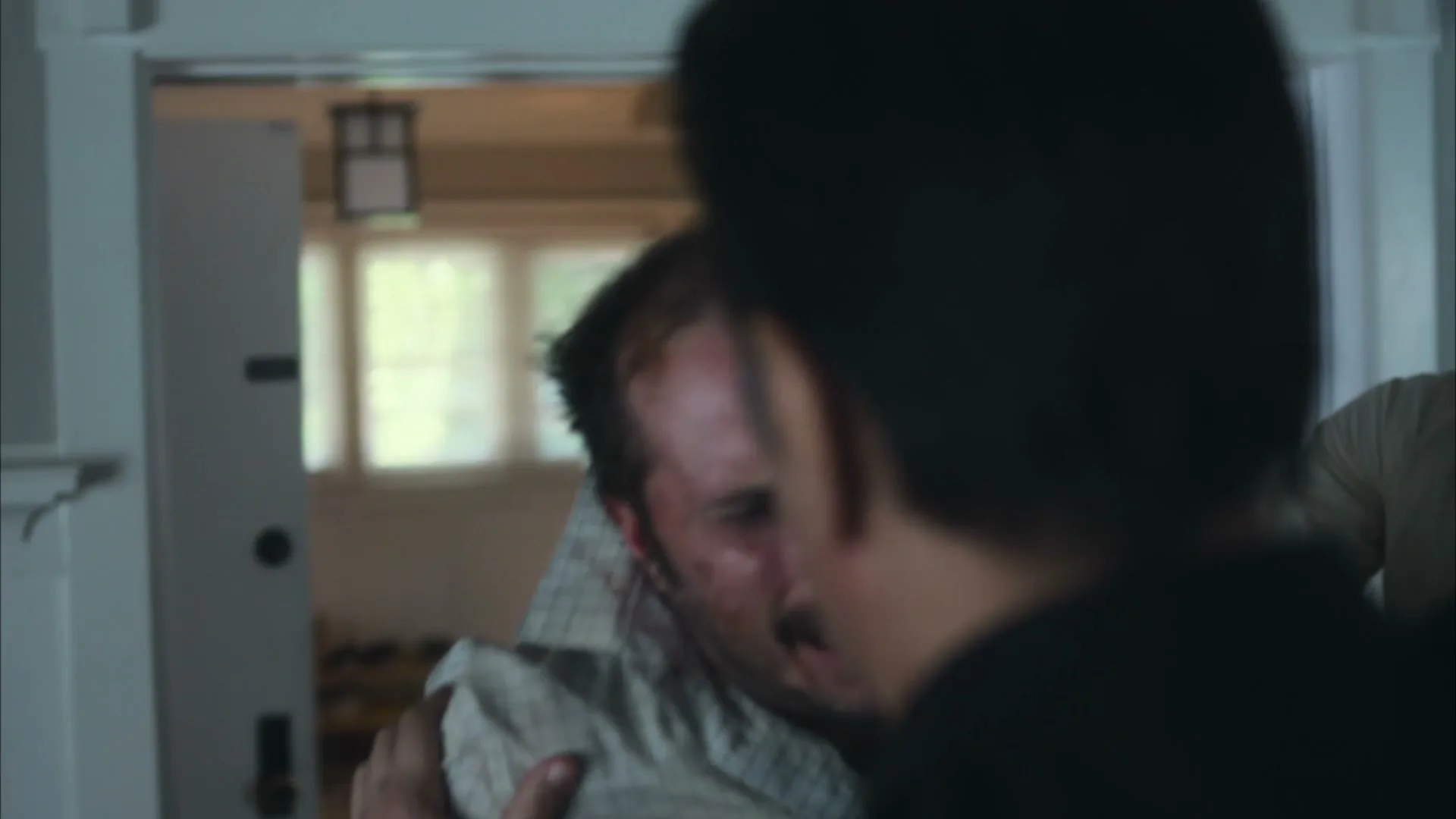 A man with a mustache and a white shirt is being held by another man in a suit, struggling in a dimly lit house.