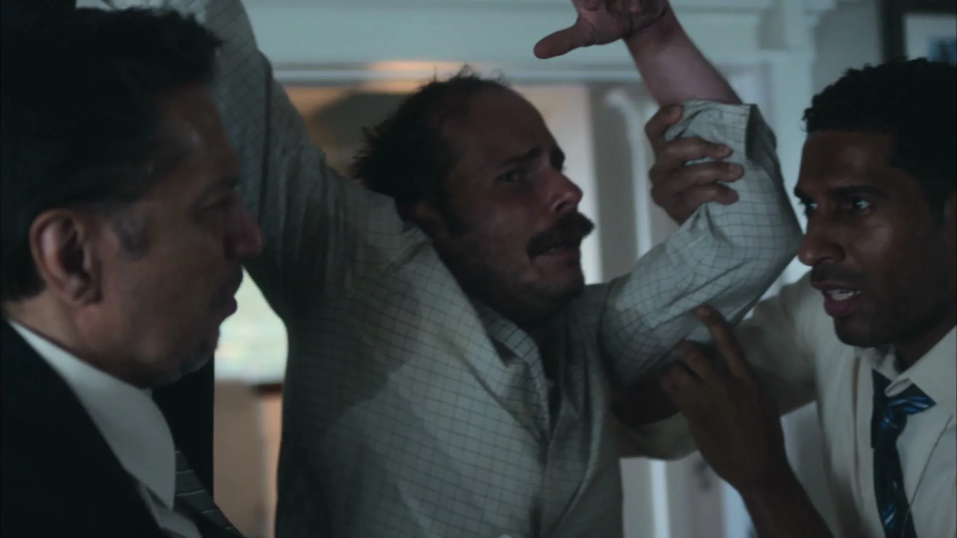Three men struggle, two holding a third man who is crying out, in a dimly lit interior setting.