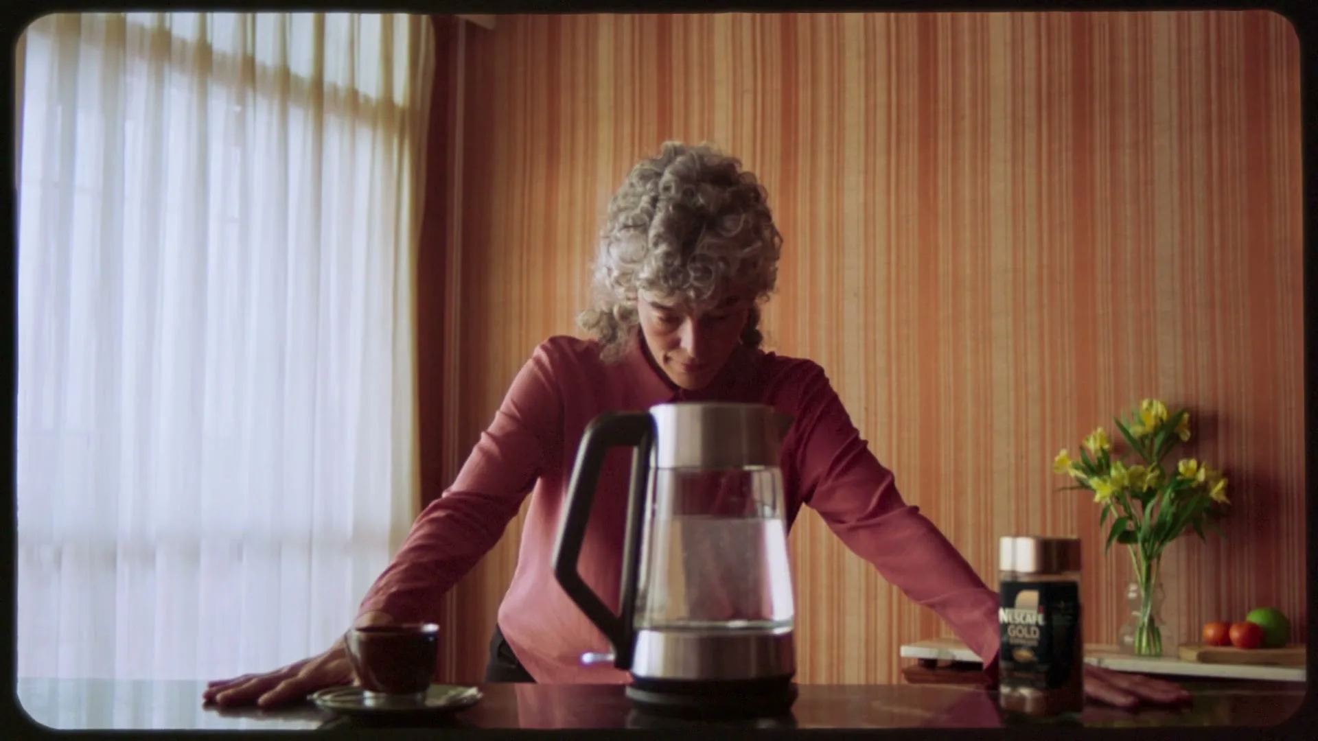 A woman with curly grey hair in a pink top leans over a kitchen counter, looking down at a kettle and a jar of Nescafé Gold.