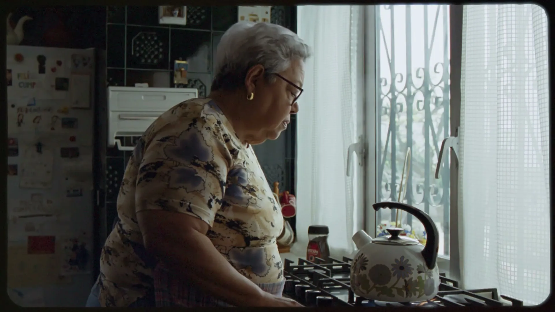 An older woman with gray hair and glasses stands in a kitchen, looking down at a kettle on a gas stove next to a window.