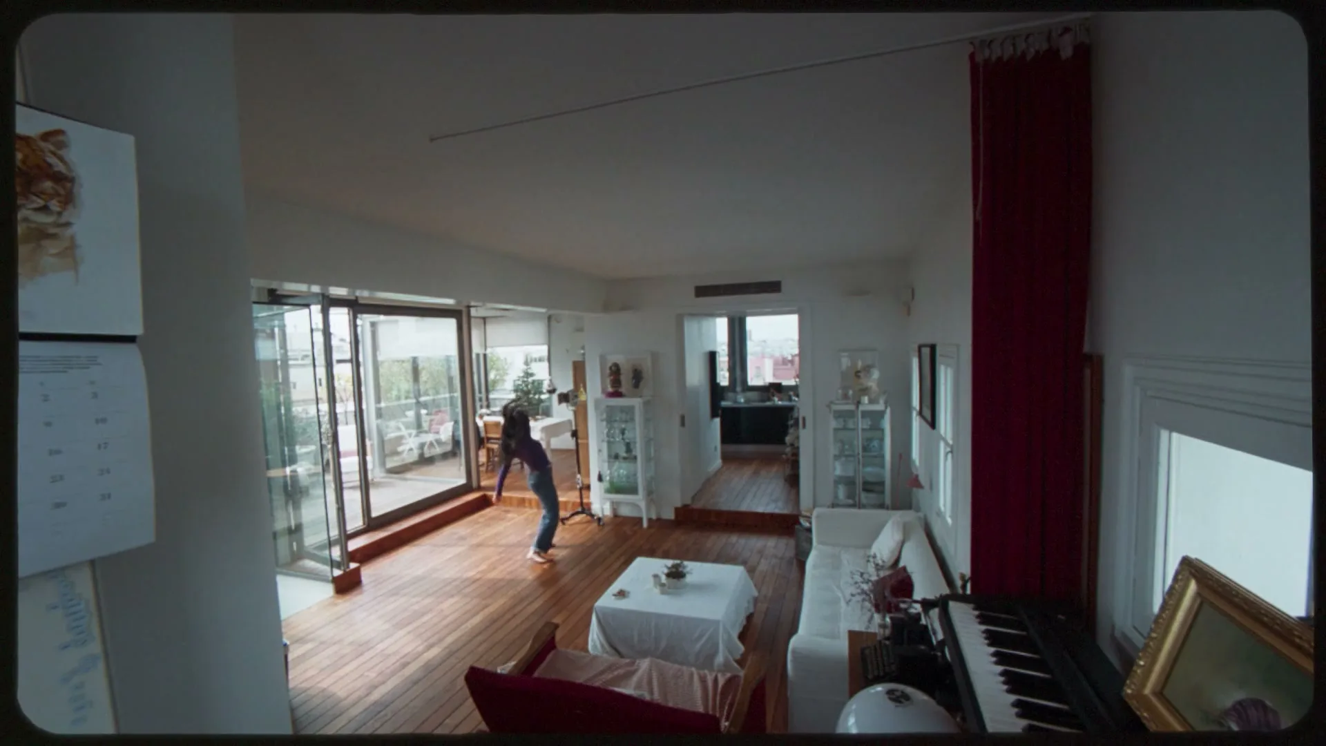 A young woman in a dark top and jeans dances playfully in a bright, modern living room with wooden floors and a balcony.