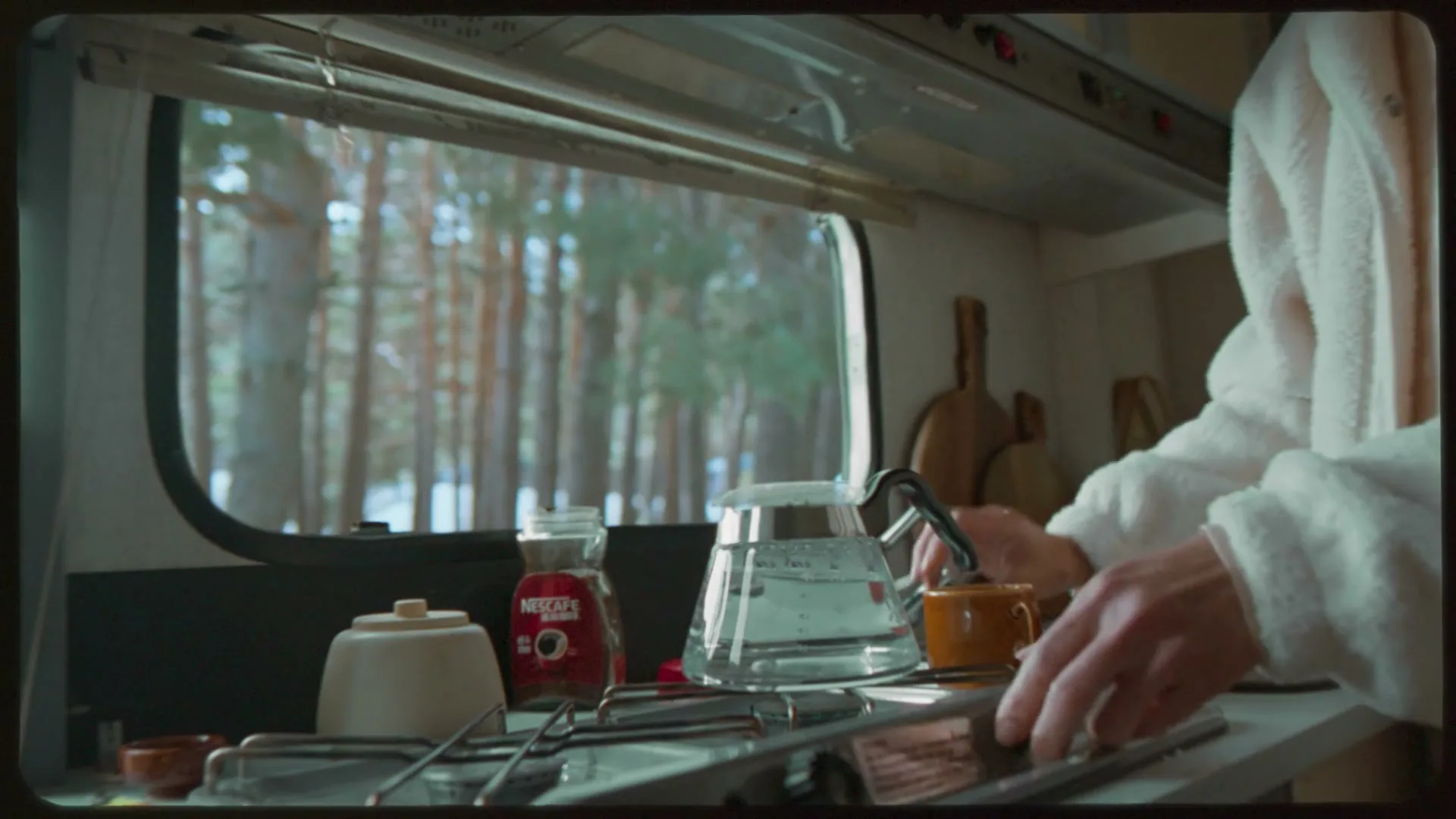 A person in a white robe stirs a cup of coffee in a camper van kitchen, with a snowy forest visible outside the window.