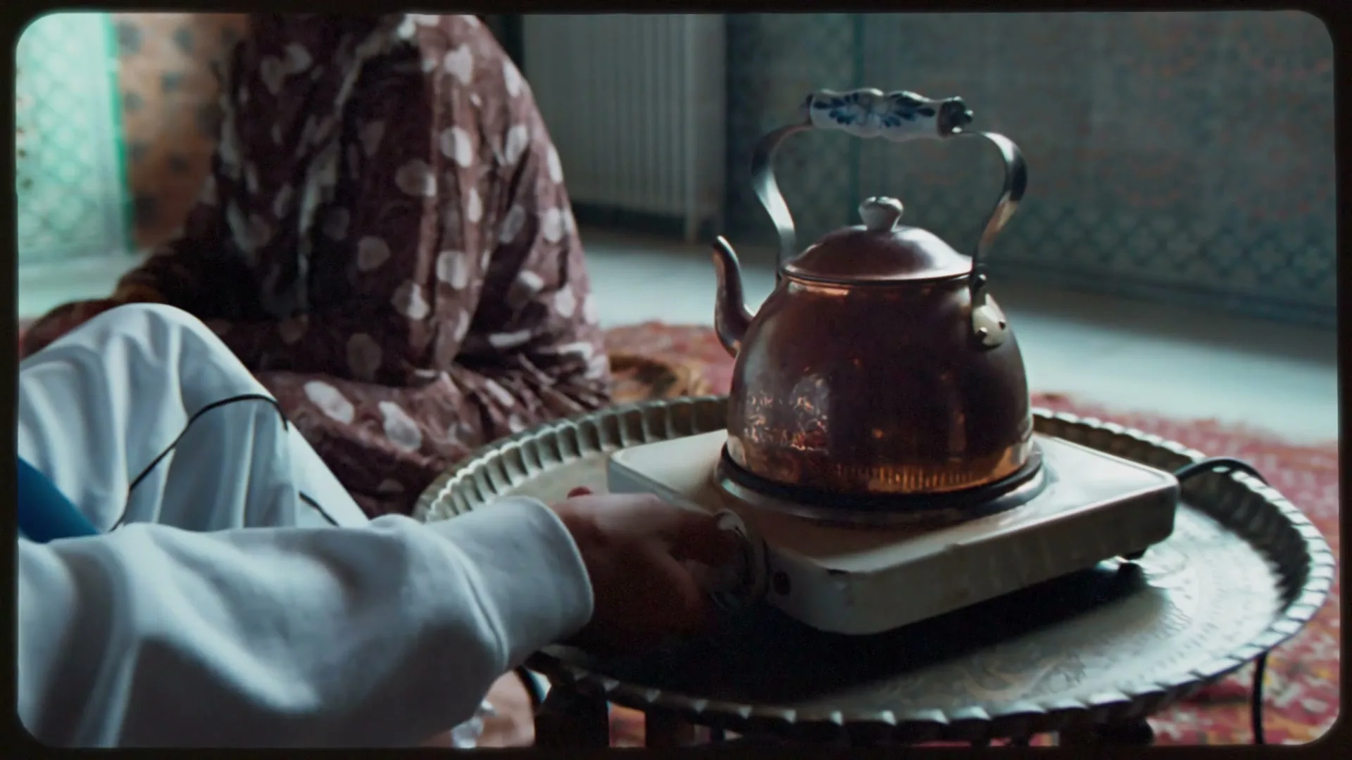 A person in a white hoodie adjusts a copper kettle on a hot plate, with another person in a patterned dress sitting in the background.