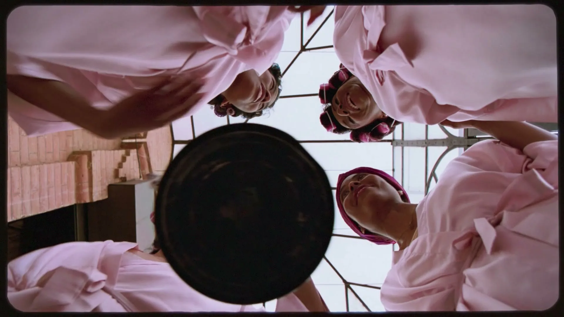 Four women in pink robes and hair curlers look down and smile at the camera, which is looking up at them from a glass ceiling.