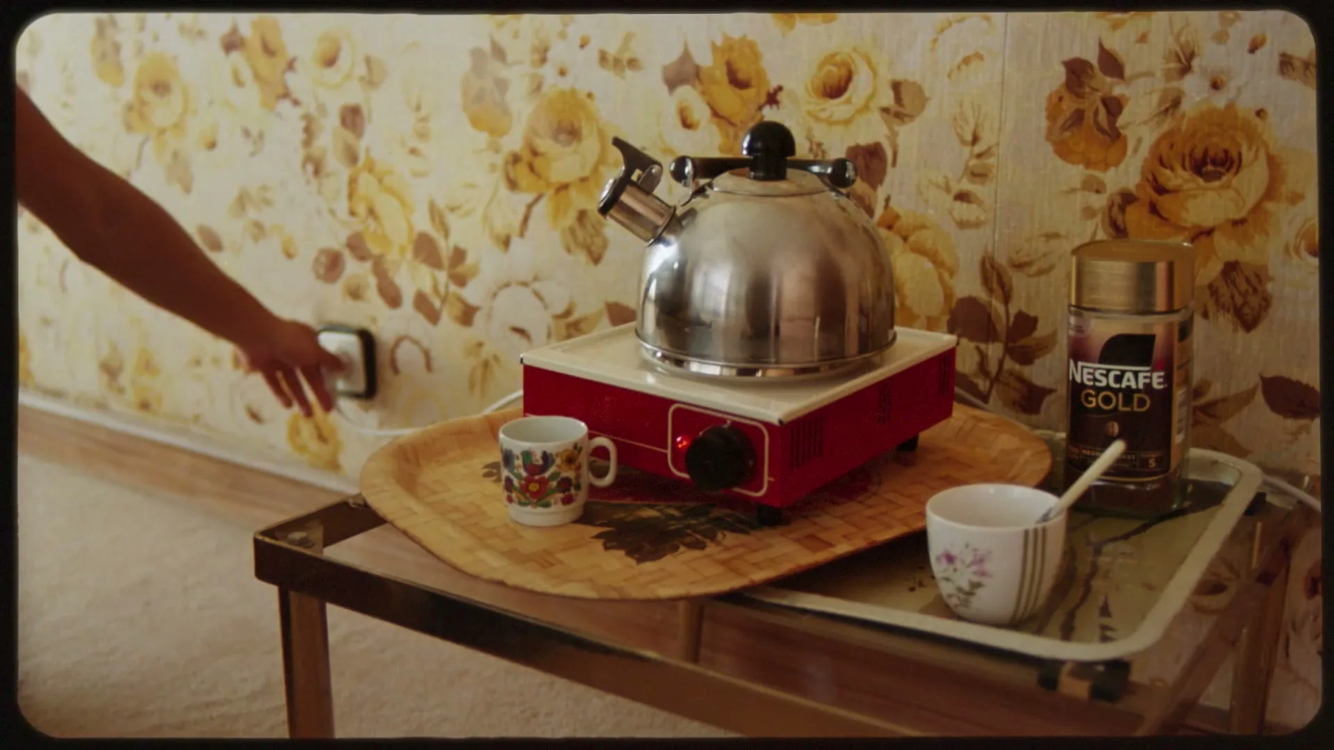 A person's arm reaches to plug in a hot plate with a kettle on it, next to a jar of Nescafé Gold, in a room with floral wallpaper.
