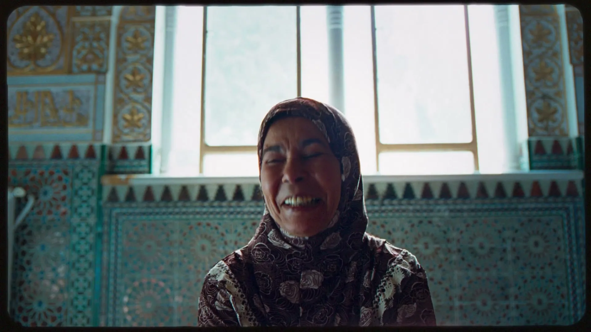 A woman in her 40s, wearing a patterned hijab, laughs heartily in a beautifully tiled room with a bright window behind her.