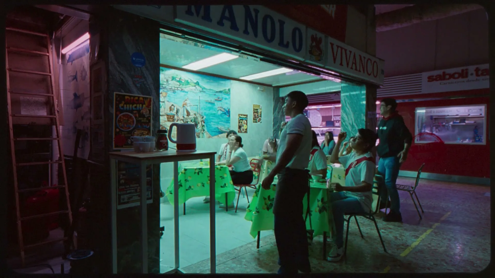 A man in a white t-shirt and apron walks past a small group of people sitting at tables in a brightly lit food stall at night.