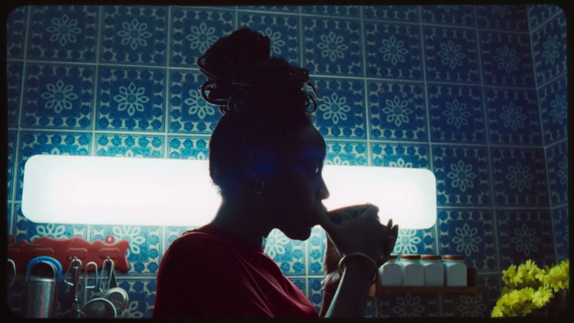 A woman in silhouette sips from a mug in a kitchen with blue patterned tiles and a bright horizontal light fixture.