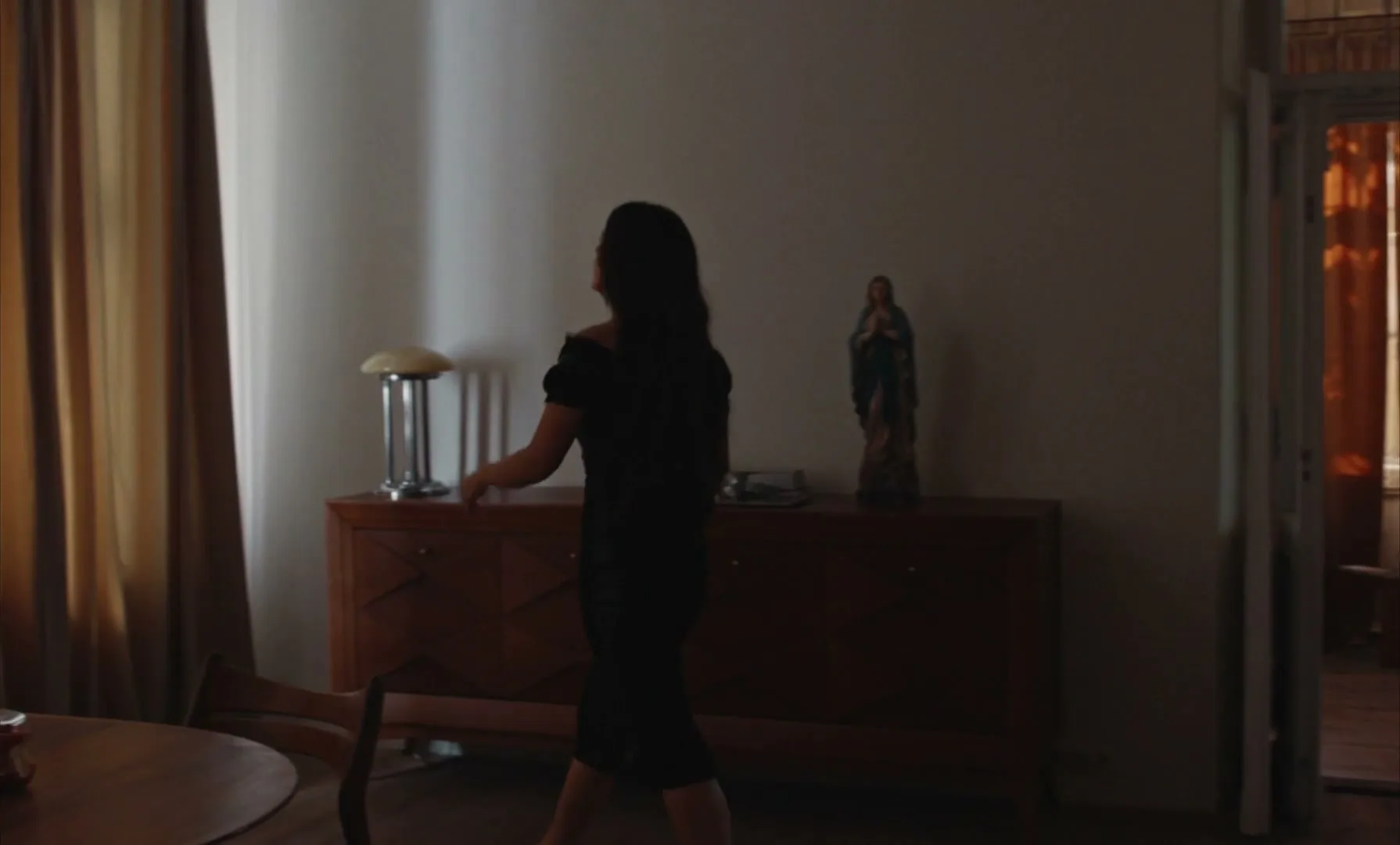 A woman in a black dress walks across a dimly lit room, then pulls back curtains to reveal a bright window.