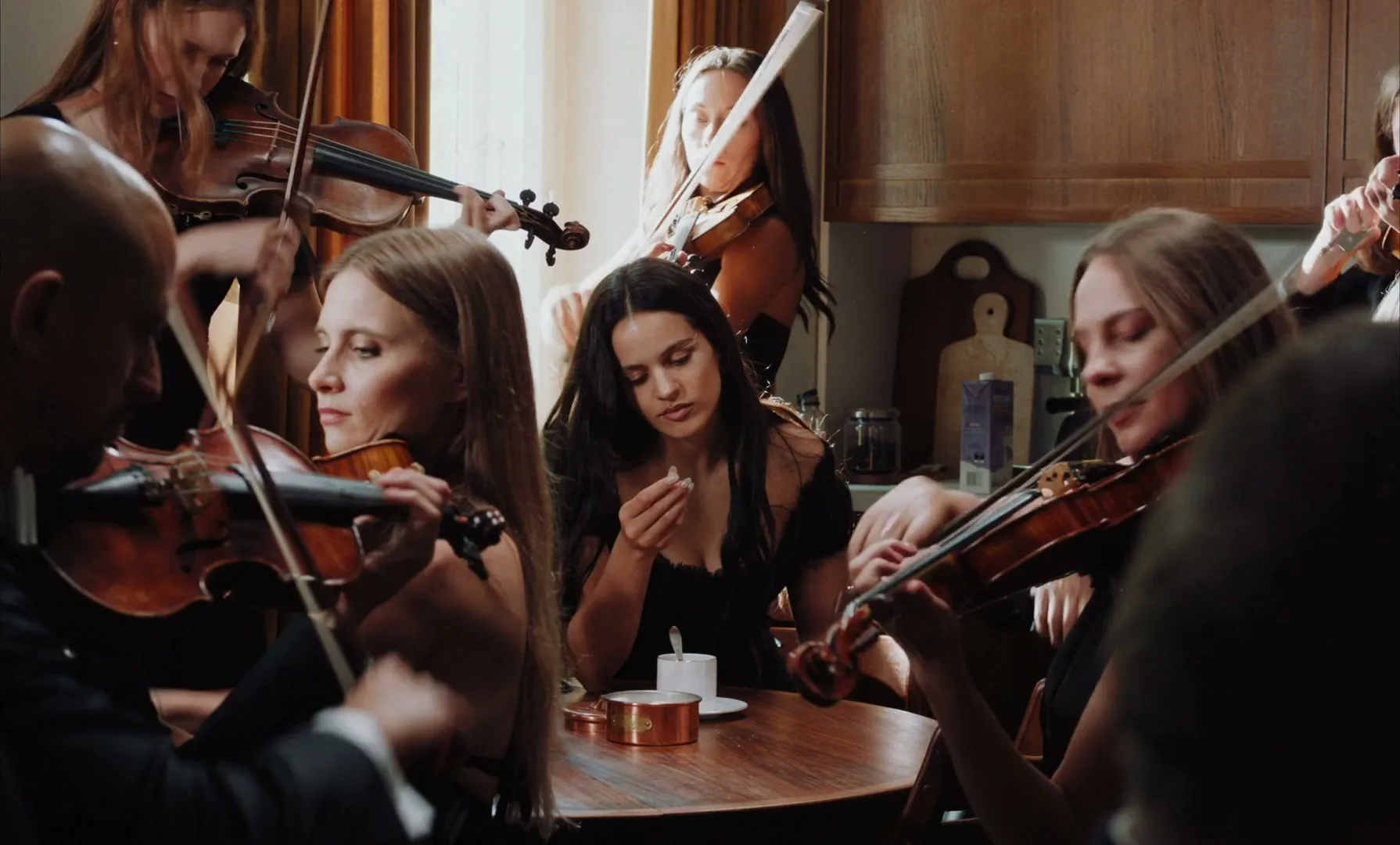 Rosalia sits at a table, looking down and eating something, surrounded by people playing violins in a warm, sunlit room.