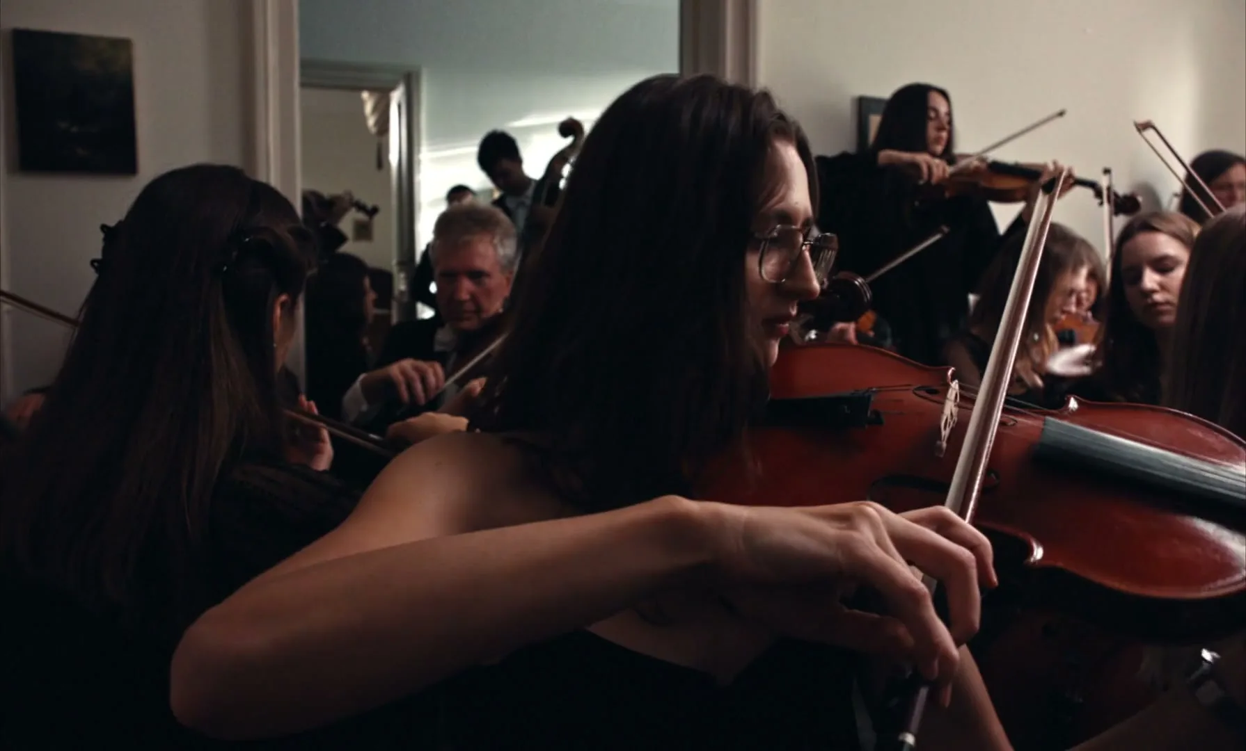 A young woman with glasses plays the violin in a crowded room with other musicians, including an older man, all dressed in dark clothing.