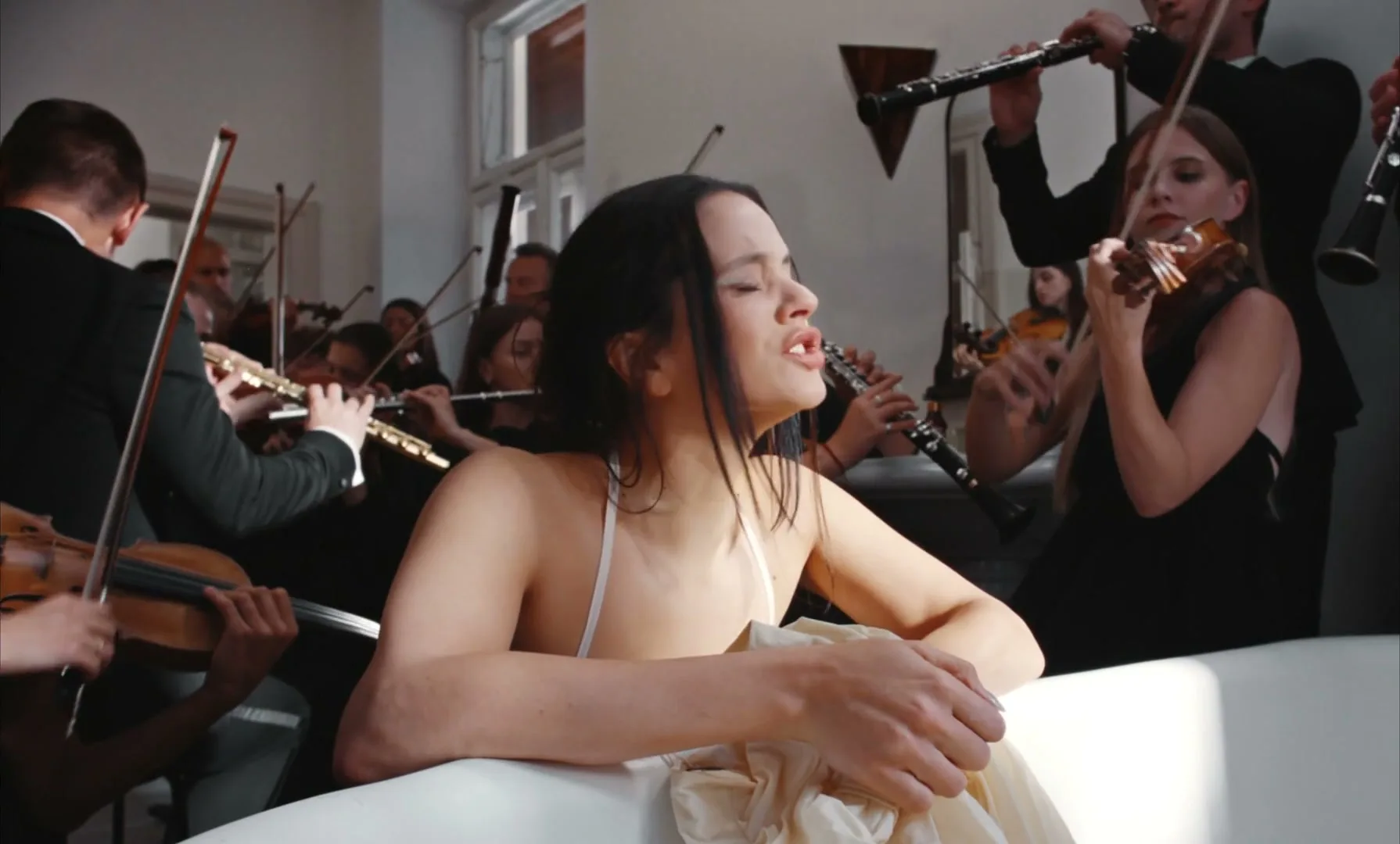 Rosalia, a young woman with dark hair, sings passionately in a bathtub surrounded by an orchestra playing classical instruments in a bright room.