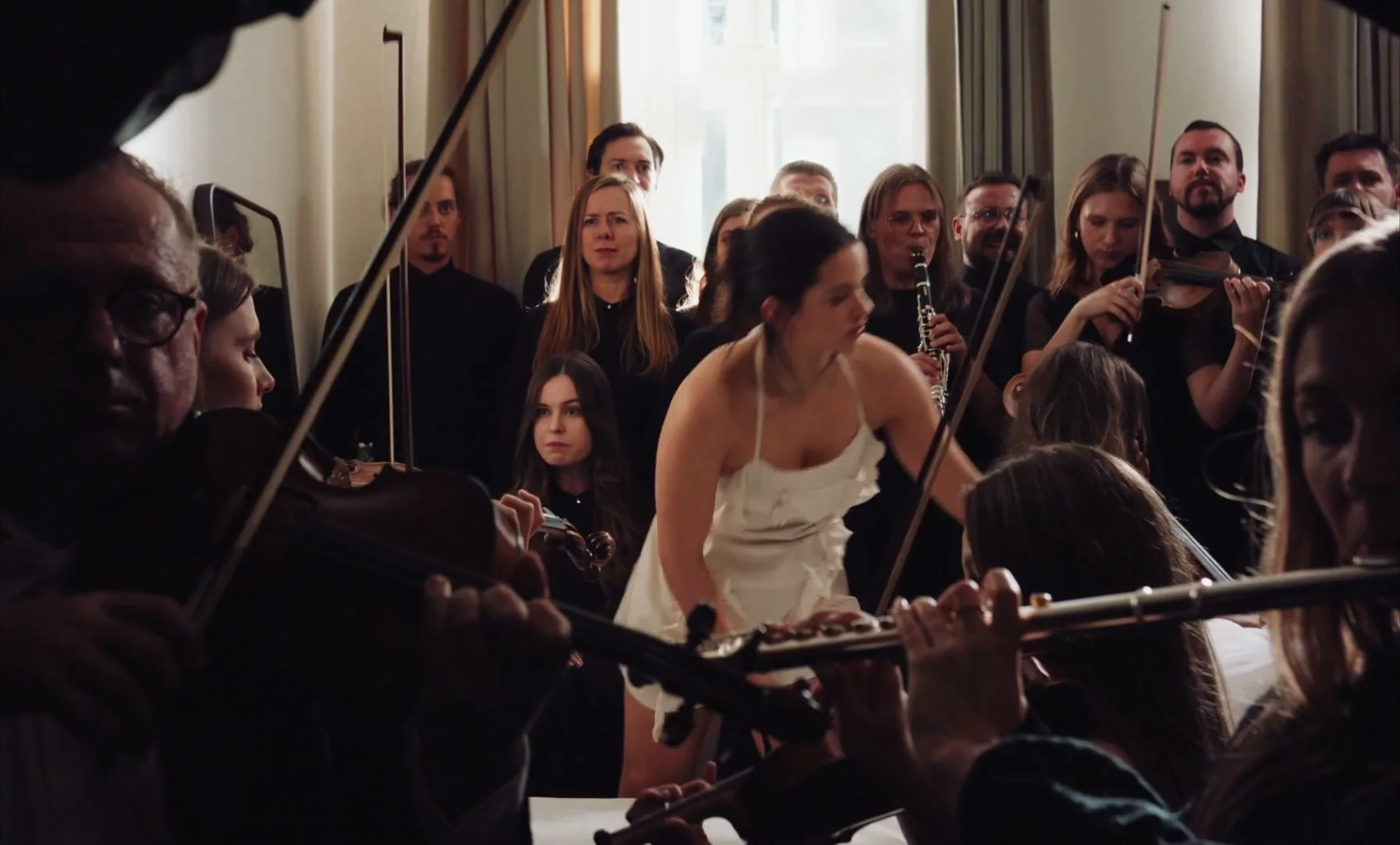 A woman in a white dress dances wildly with a pillow in a room full of formally dressed orchestra members playing instruments.