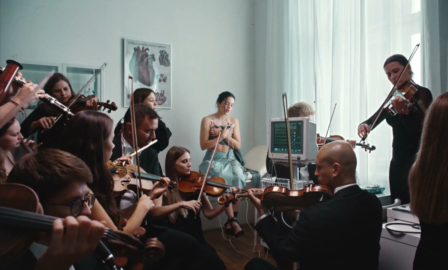 Rosal

ia sits in a hospital gown with an ECG machine attached, surrounded by an orchestra playing string instruments in a bright room.