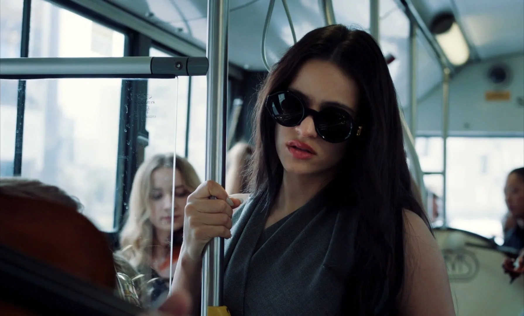 Rosalía, a young woman with long dark hair and sunglasses, holds a pole on a moving bus, looking around with a slightly open mouth.