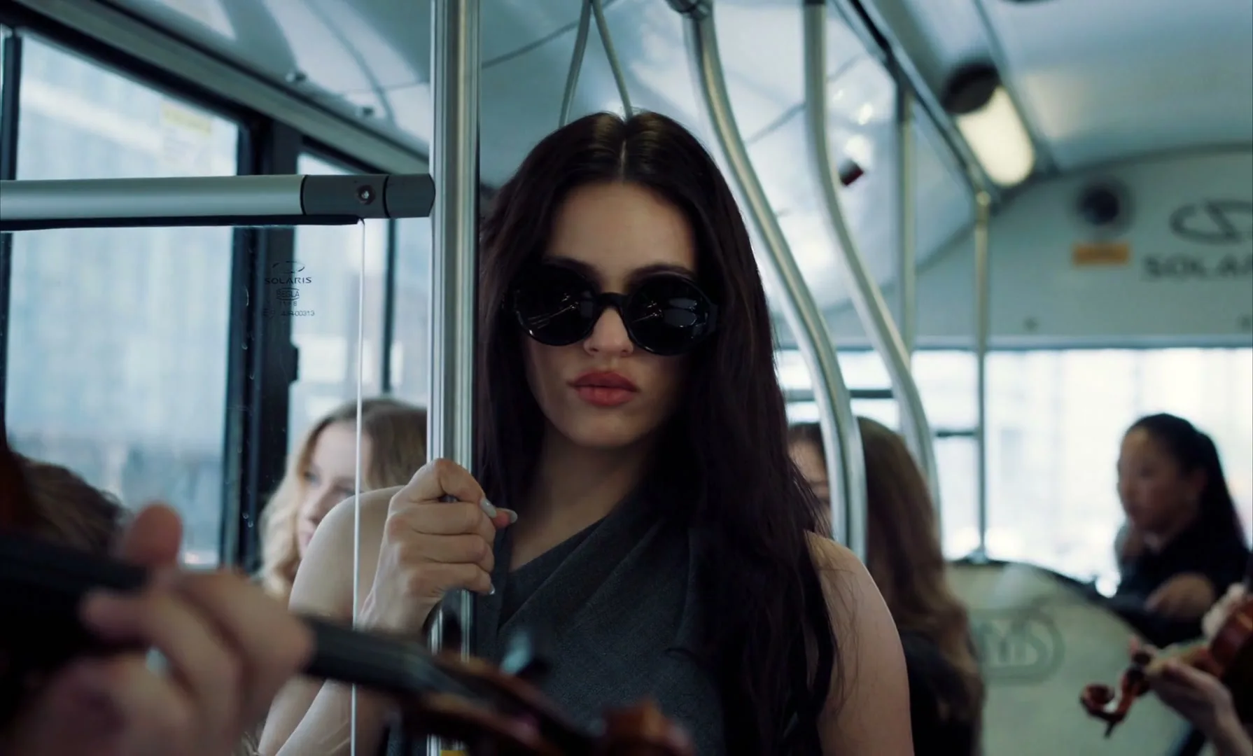 A young woman with long dark hair and sunglasses stands on a bus, holding a pole, with other passengers and musicians around her.