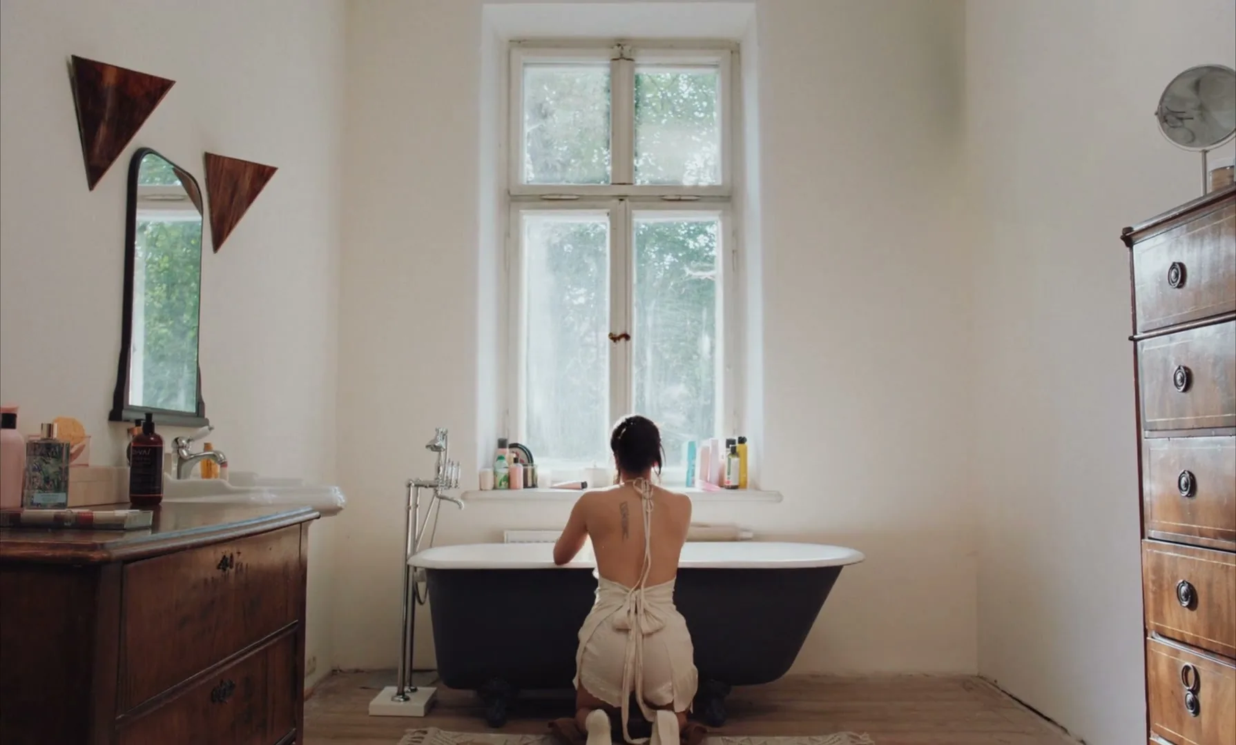 A woman with dark hair, wearing a white dress, kneels in front of a bathtub, cleaning it in a bright, minimalist bathroom.