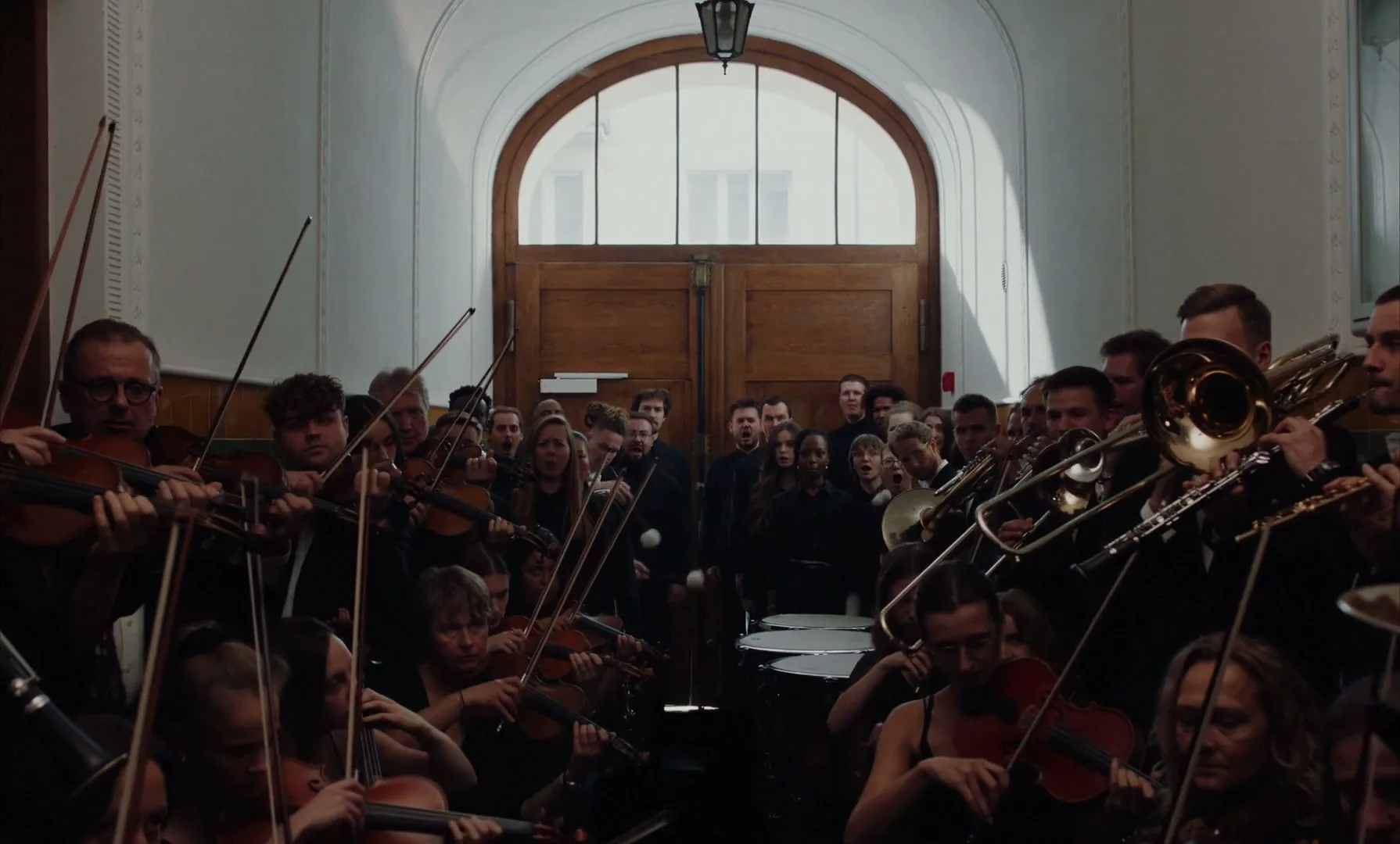 A large orchestra of diverse people in black attire plays their instruments in a grand hall with a large arched window and wooden doors.