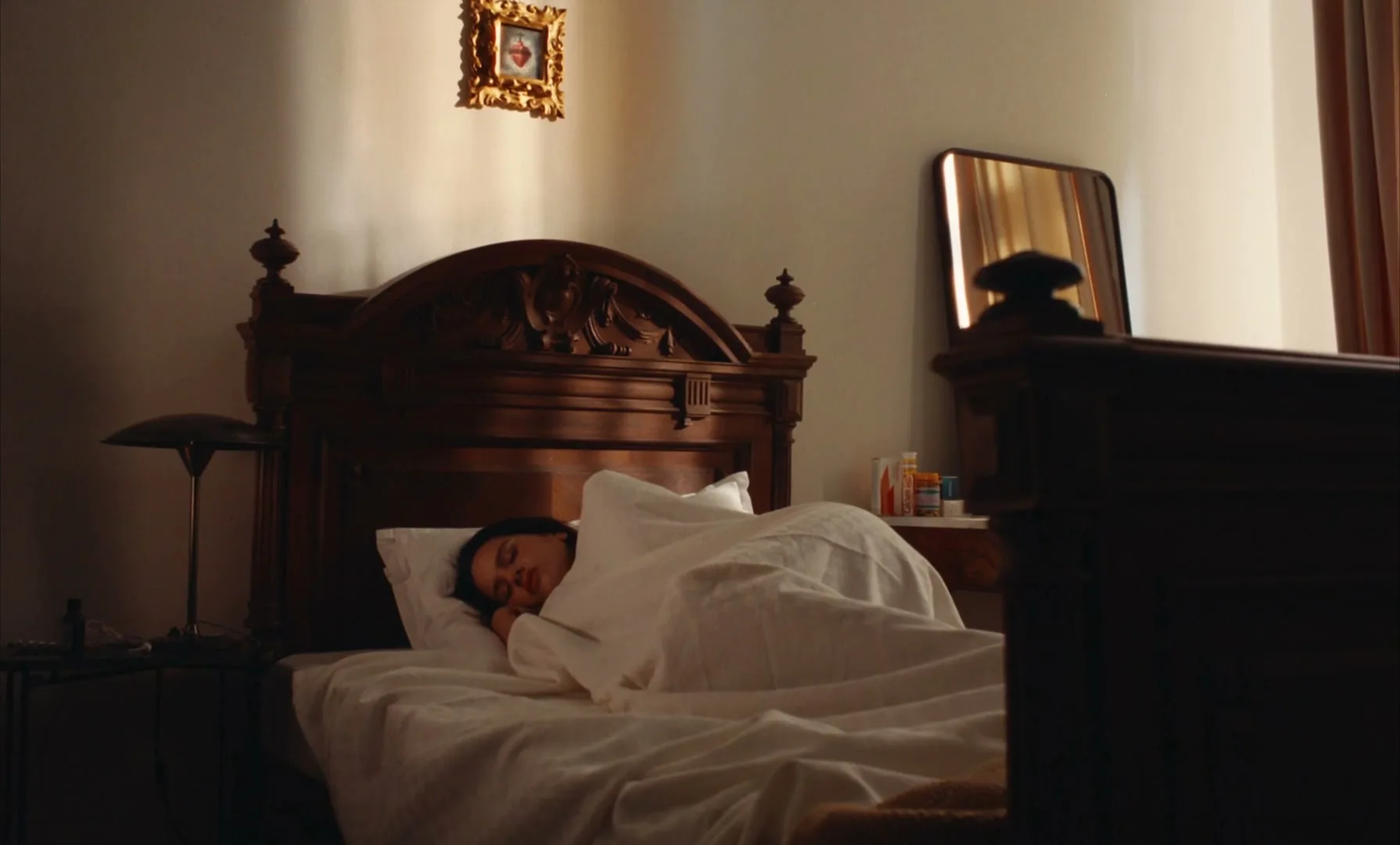 A young woman with dark hair sleeps peacefully under white sheets in a dimly lit bedroom with antique wooden furniture.