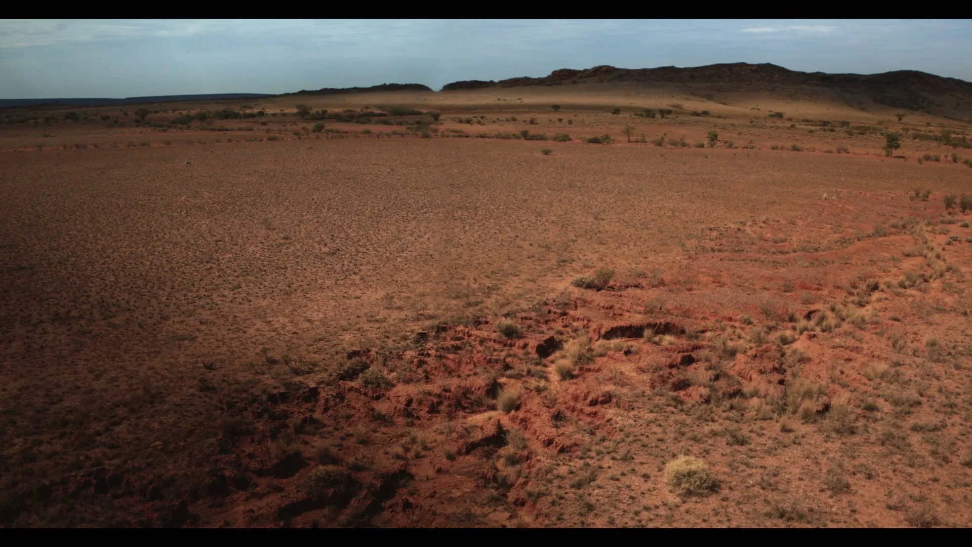 An aerial wide shot captures a vast, arid landscape with reddish-brown earth and sparse vegetation under a partly cloudy sky, with text overlay.
