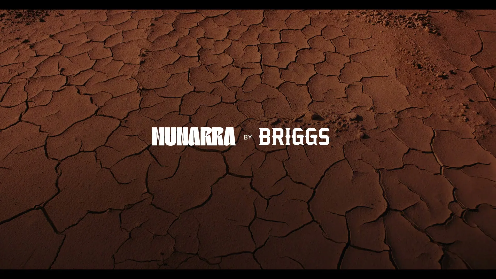The camera looks down at a dry, cracked desert floor with the words "MUNARRA by BRIGGS" in white text across the middle.