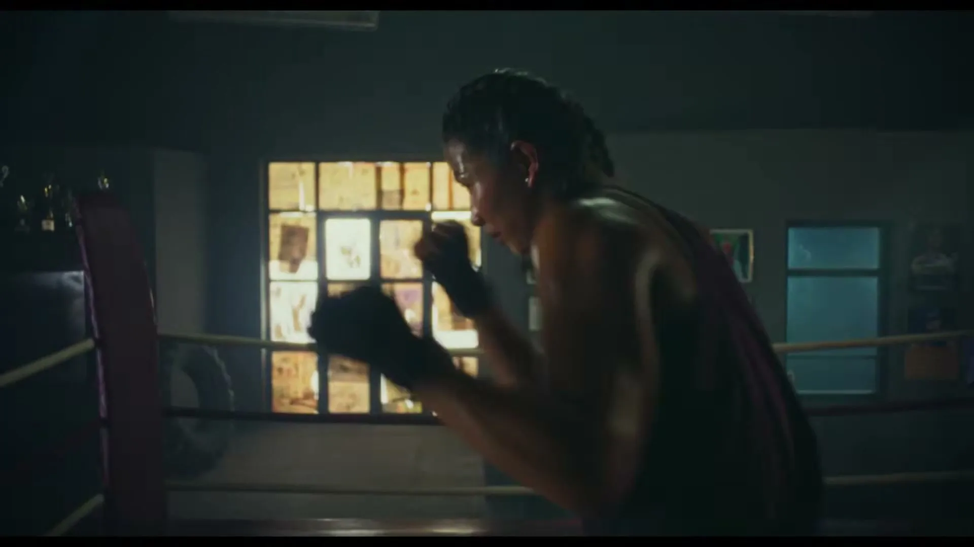 A female boxer with braided hair, wearing a purple tank top and black hand wraps, trains in a boxing ring in a dimly lit gym.