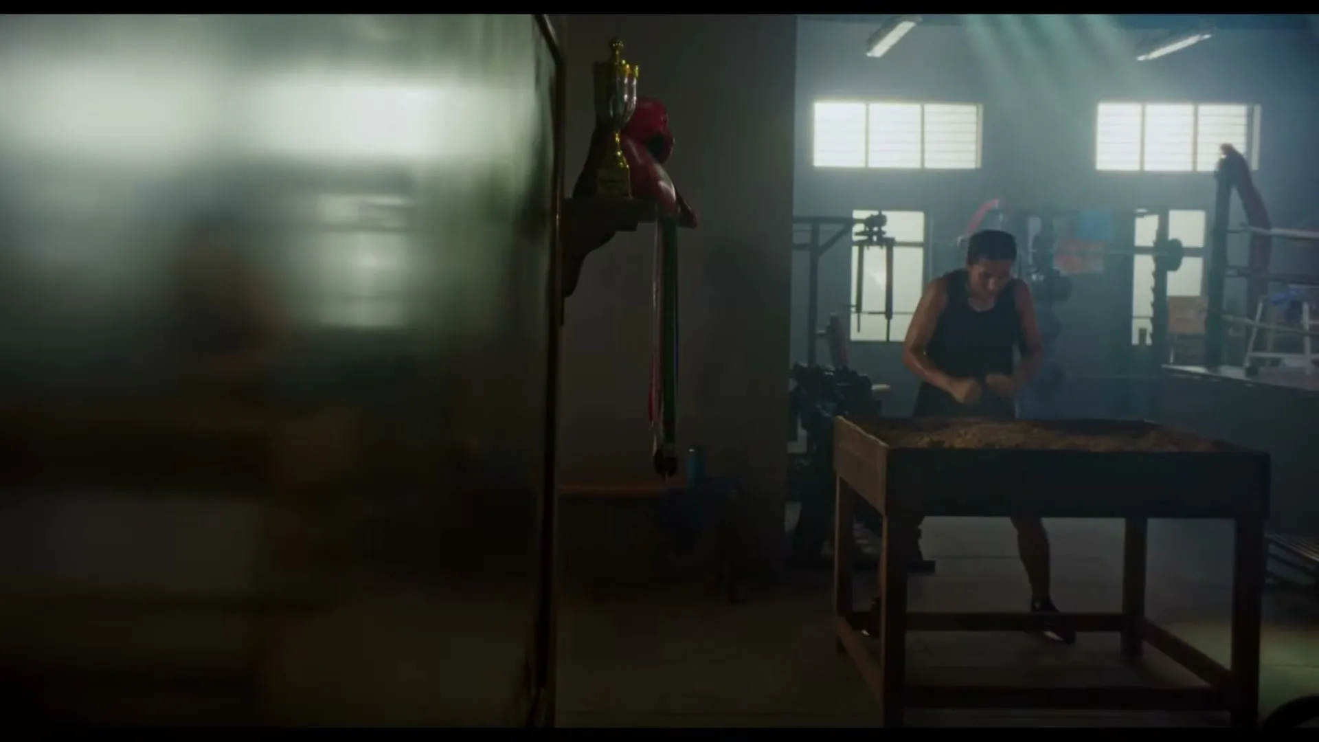 A female boxer in a black tank top trains by punching sand in a wooden box in a dimly lit gym.