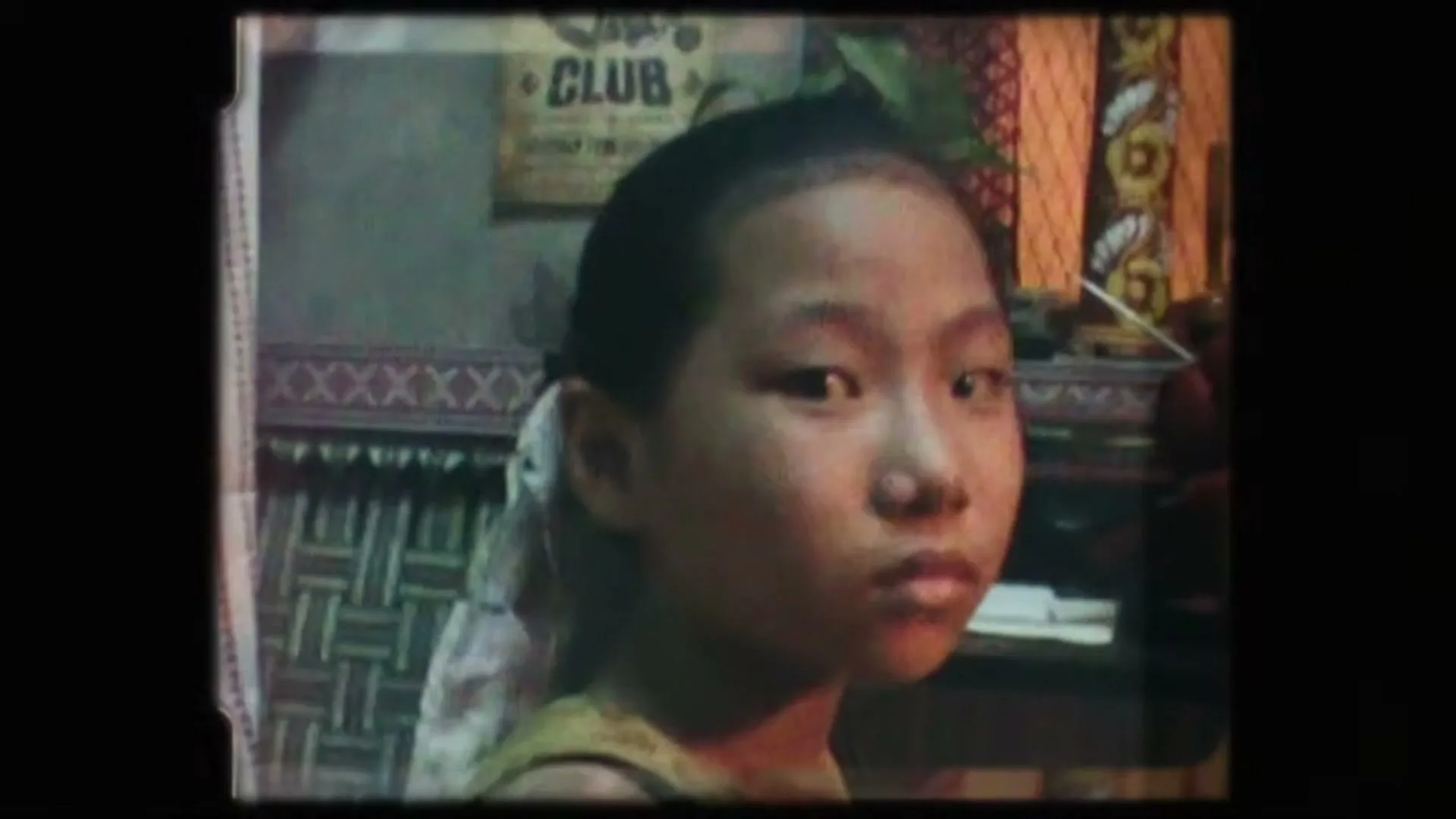 A young girl with dark hair pulled back looks directly at the camera with a neutral expression in a dimly lit room.