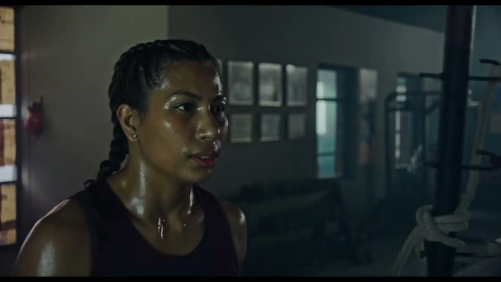 A female boxer, Lovelina Borgohain, with braided hair and a sweaty face, looks intently to the side in a dimly lit boxing gym.