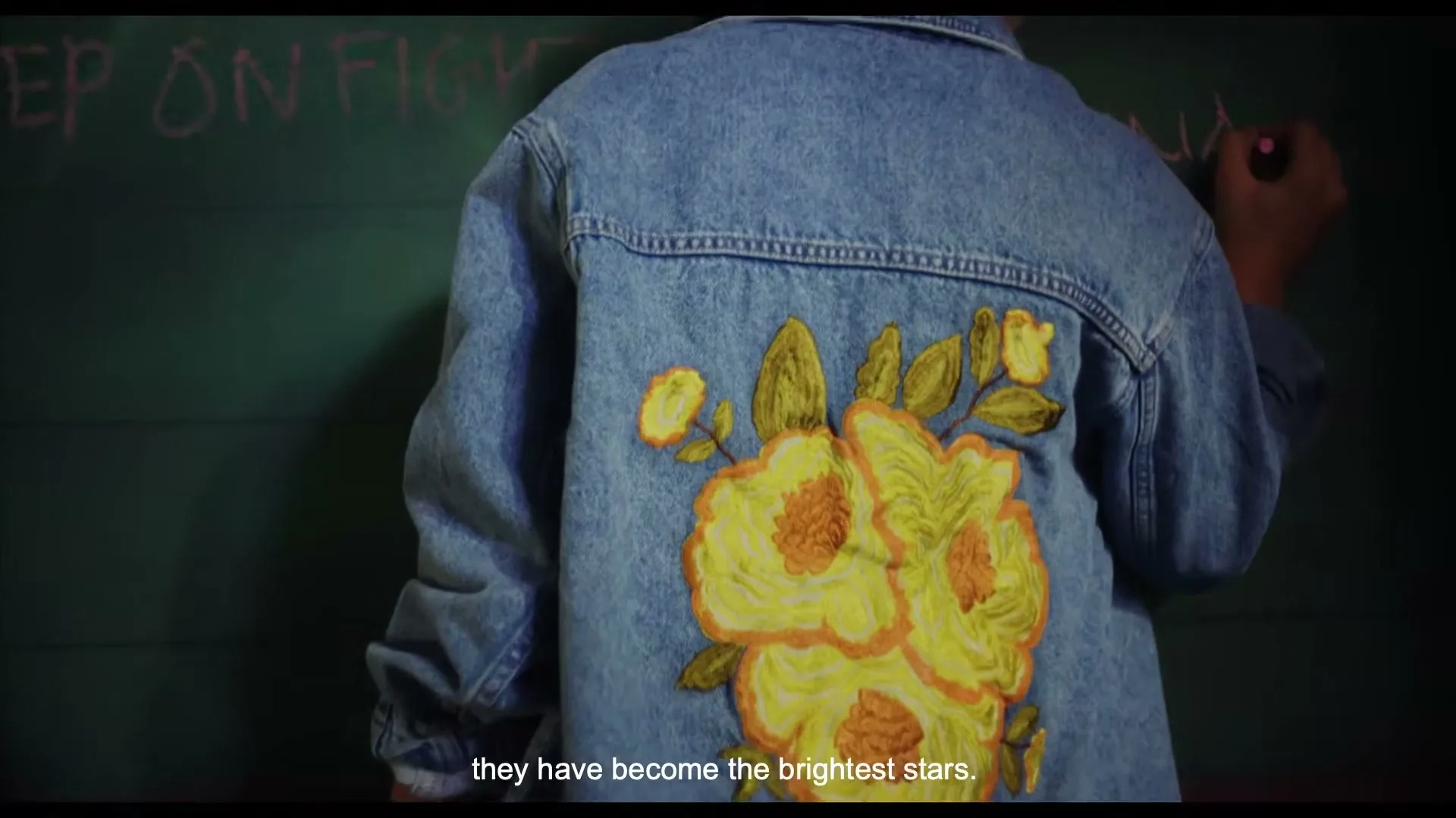 A person in a denim jacket with yellow floral embroidery writes on a green chalkboard, seen from behind in a medium shot.