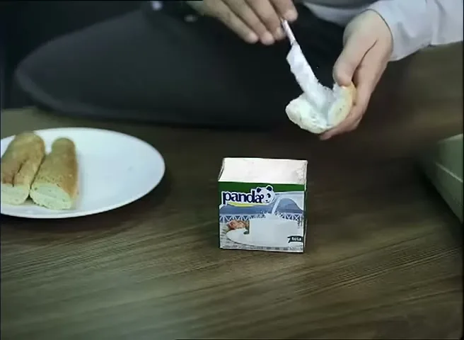 A person's hands spread a white creamy substance from a small carton of Panda cheese onto a piece of bread on a wooden floor.