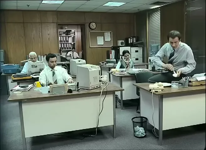 An office worker in a white shirt and tie sits on a desk, eating from a small bowl, while other office workers are busy at their desks in a retro office.