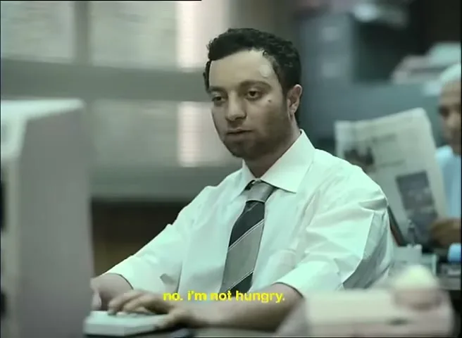 A man with dark hair and a white shirt works at a computer in a busy office, looking tired and slightly annoyed.