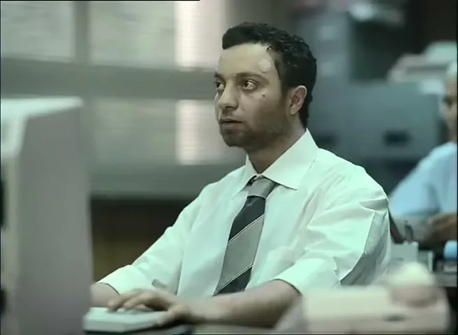 A man with a bandage on his forehead sits at a computer in a busy office, looking tired and stressed.