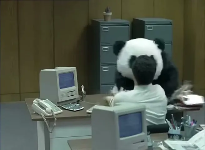 A man in a white shirt sits at a desk, looking at a panda mascot who is standing over him in an office.