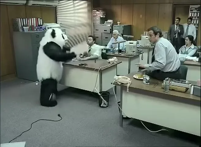 A person in a panda suit smashes a computer monitor on a desk in an office, while office workers look on in shock.